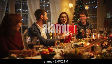 Eltern und Verwandte genießen am Abend gemeinsam ein Weihnachtsessen in einem gemütlichen Heim. Schöne Freunde versammelten sich, um den Geburtstag einer jungen Frau zu feiern. Glücklicher Mann, der ein festliches Geschenk überreicht Stockfoto