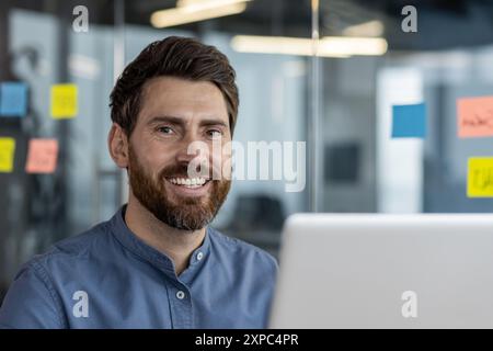 Der lächelnde Mann arbeitet an einem Laptop in einer modernen Büroumgebung mit Glaswänden und farbenfrohen Haftnotizen. Der Mann zeigt positive Einstellung und Professionalität. Stockfoto