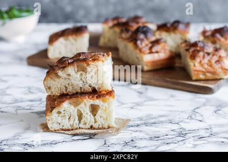 Nahaufnahme von zwei Scheiben goldenem, luftigem Rosmarin-Focaccia-Brot, gestapelt auf einer Marmoroberfläche, mit weiteren Stücken im Hintergrund auf einem Holzbrett Stockfoto