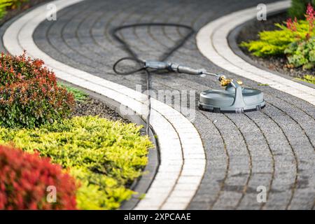 Ein Hochdruckreiniger befindet sich auf einem geschwungenen Gartenweg. An den Seiten flankieren leuchtende Sträucher und Pflanzen, was darauf hinweist, dass gerade Wartungsarbeiten durchgeführt werden. Stockfoto