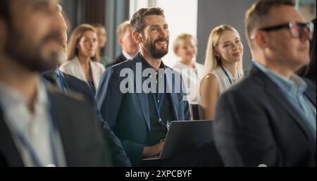 Ein kaukasischer Mann sitzt in einem überfüllten Publikum auf einer IT-Konferenz. Teilnehmer verwenden Laptop-Computer, um Notizen zu machen. Softwareentwickler sehen sich die Präsentation über neue High-Tech-Produkte und Cloud-Lösungen an Stockfoto