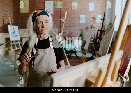 Moderne ältere kaukasische Frau mit Palette und Pinselmalerei auf Leinwand im Kunststudio Stockfoto