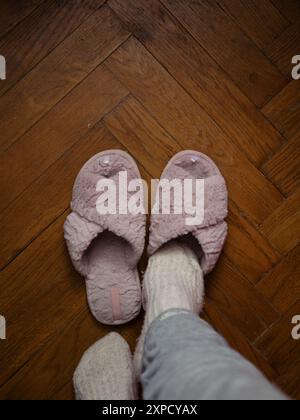 Direkt über der vertikalen Nahaufnahme einer Frau in kuscheligen Schlafanzügen treten weiße Wollsocken in ihre rosa flauschigen Hausschuhe auf einem Parkettboden. Offenes Zuhause Stockfoto