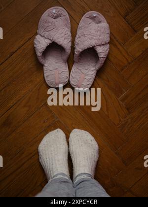 Direkt über der vertikalen Nahaufnahme einer Frau, die in weißen Woollsocken auf einem Parkettboden vor den rosa flauschigen Hausschuhen steht. Freimütige Heimaufnahme. Stockfoto