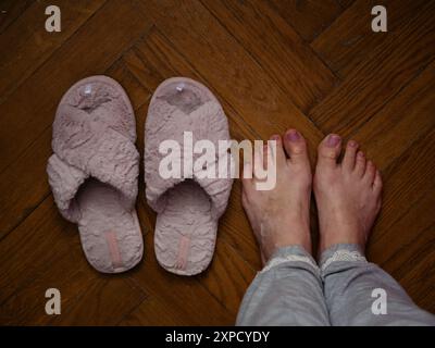 Direkt über der horizontalen Nahaufnahme einer Frau im grauen Pyjama, die barfuß auf einem Parkettboden neben den rosa flauschigen Hausschuhen mit Pedicure steht Stockfoto
