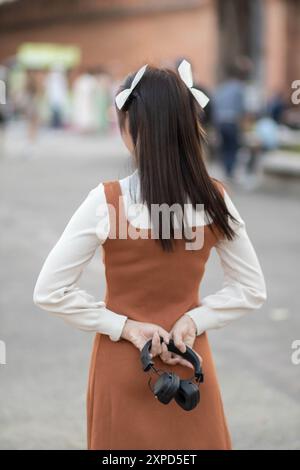 Hinter den Kulissen steht eine junge Frau im Vintage-Stil, die das Bluetooth-Headset in der Hand hält, und geht allein mit Musikkopfhörern Stockfoto