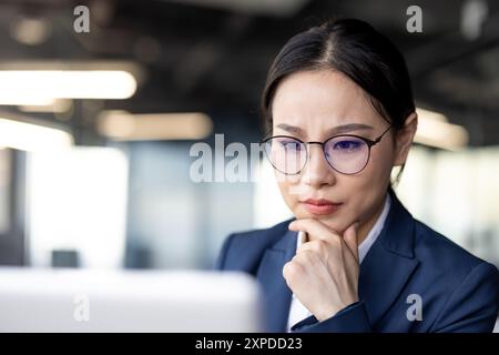 Eine fokussierte asiatische Geschäftsfrau, die an ihrem Laptop in einer modernen Büroumgebung arbeitet. Sie trägt einen marineblauen Anzug und eine Brille, konzentriert sich auf ihre Aufgaben und zeigt Entschlossenheit und Professionalität. Stockfoto