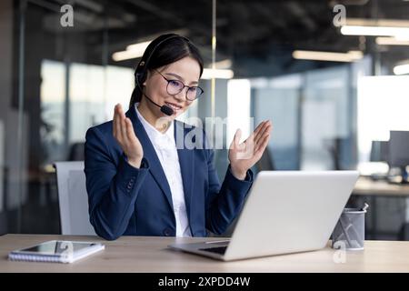 Professionelle asiatische Geschäftsfrau in einer modernen Büroumgebung, die ein Headset trägt und einen Videoanruf auf ihrem Laptop führt. Sie wirkt engagiert und selbstbewusst und sitzt mit einem Notizbuch an einem Holzschreibtisch Stockfoto