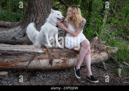 Eine blonde Frau in einem weißen Kleid sitzt auf einem Holzstamm mit einem weißen Hund in der Nähe eines Willamette-Flusses Stockfoto