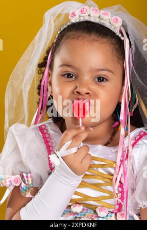 Ein Mädchen als Braut gekleidet, fröhlich und schön, mit einem roten Lutscher im Mund. Porträt mit gelbem Hintergrund im Studio. Stockfoto