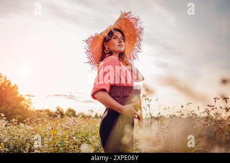 Porträt einer jungen, stilvollen Frau, die auf einem blühenden Buckweat-Feld mit Strohhut und Ledertasche läuft Stockfoto