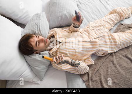 Junger Mann im Schlafanzug mit elektronischer Wegwerfzigarette mit Handy auf dem Bett zu Hause Stockfoto