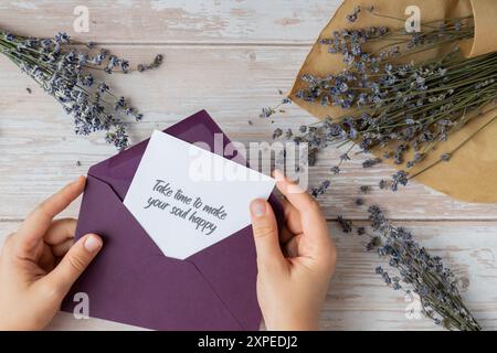 Weibliche Hände, die Papierkarten mit Text nehmen, NEHMEN SICH ZEIT, UM IHRE SEELE mit violettem Umschlag GLÜCKLICH ZU MACHEN. Lavendelblume. Draufsicht, flach. Konzept der psychisch-spirituellen Gesundheit Selbstversorgung Wohlbefinden Achtsamkeit Stockfoto