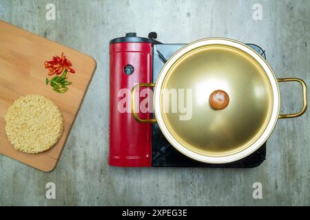 Ein roter Topf mit goldenem Deckel sitzt auf einem Brenner. Neben dem Topf befindet sich ein hölzernes Schneidebrett mit einer Schüssel Instant-Nudeln und einem Haufen Paprika Stockfoto