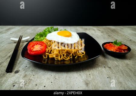 Ein Teller mit Instantnudeln mit einem Ei und einer Tomatenscheibe. Der Teller liegt auf einem Tisch mit Essstäbchen und einer Schüssel Soße Stockfoto