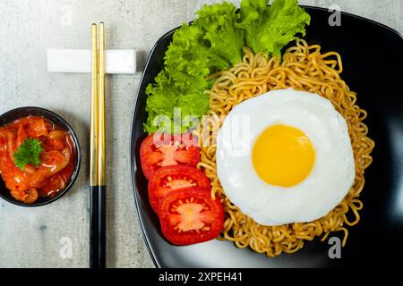 Ein Teller mit Instantnudeln mit einem Ei und einer Tomatenscheibe. Der Teller liegt auf einem Tisch mit Essstäbchen und einer Schüssel Soße Stockfoto