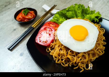 Ein Teller mit Instantnudeln mit einem Ei und einer Tomatenscheibe. Der Teller liegt auf einem Tisch mit Essstäbchen und einer Schüssel Soße Stockfoto