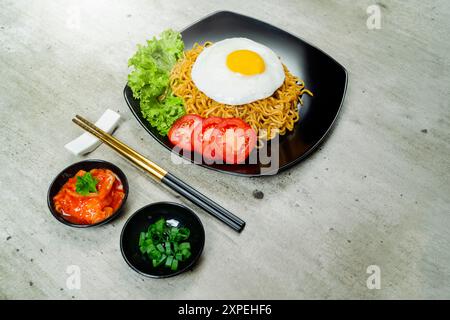 Ein Teller mit Instantnudeln mit einem Ei und einer Tomatenscheibe. Der Teller liegt auf einem Tisch mit Essstäbchen und einer Schüssel Soße Stockfoto