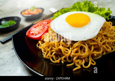 Ein Teller mit Instantnudeln mit einem Ei und einer Tomatenscheibe. Der Teller liegt auf einem Tisch mit Essstäbchen und einer Schüssel Soße Stockfoto