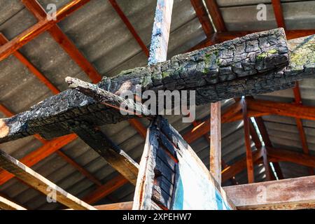 Verkohlter Holzstamm hängt von der Decke eines Gebäudes. Verkohltes Holz hat ein dunkles, rauchiges Aussehen und ist von einem grauen Dach umgeben. Konzept der Zerstörung Stockfoto