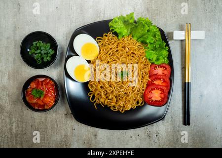 Ein Teller mit Instantnudeln mit Tomaten und Ei darauf. Der Teller wird auf einen Tisch mit Essstäbchen und Schüsseln gestellt Stockfoto