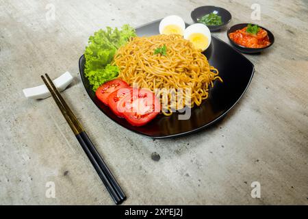 Ein Teller mit Instantnudeln mit Tomaten und Ei darauf. Der Teller wird auf einen Tisch mit Essstäbchen und Schüsseln gestellt Stockfoto
