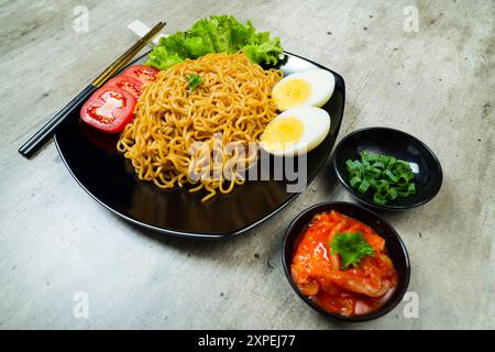 Ein Teller mit Instantnudeln mit Tomaten und Ei darauf. Der Teller wird auf einen Tisch mit Essstäbchen und Schüsseln gestellt Stockfoto