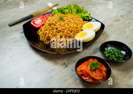 Ein Teller mit Instantnudeln mit Tomaten und Ei darauf. Der Teller wird auf einen Tisch mit Essstäbchen und Schüsseln gestellt Stockfoto