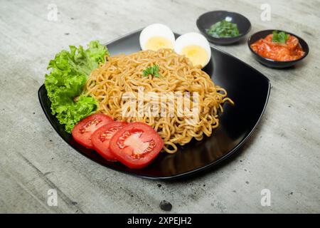 Ein Teller mit Instantnudeln mit Tomaten und Ei darauf. Der Teller wird auf einen Tisch mit Essstäbchen und Schüsseln gestellt Stockfoto