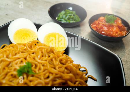 Ein Teller mit Instantnudeln mit Tomaten und Ei darauf. Der Teller wird auf einen Tisch mit Essstäbchen und Schüsseln gestellt Stockfoto