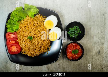 Ein Teller mit Instantnudeln mit Tomaten und Ei darauf. Der Teller wird auf einen Tisch mit Essstäbchen und Schüsseln gestellt Stockfoto