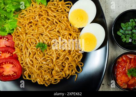 Ein Teller mit Instantnudeln mit Tomaten und Ei darauf. Der Teller wird auf einen Tisch mit Essstäbchen und Schüsseln gestellt Stockfoto