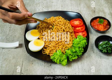 Eine Person isst einen Teller mit Instant-Nudeln mit Eiern und Tomaten. Der Teller liegt auf einem Tisch mit einer Schüssel Soße und einem Löffel. Die Szene ist lässig und re Stockfoto