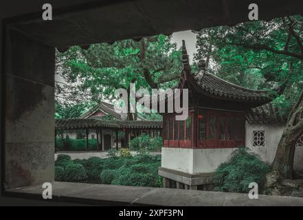 Ein Blick durch ein Fenster in einen traditionellen chinesischen Garten mit einem hölzernen Pavillon und üppigem Grün. Stockfoto
