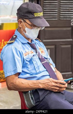 Ein Wächter in Uniform sitzt auf einem Stuhl am Bankeingang in Hoi an Vietnam. Der vietnamesische Sicherheitsbeamte sitzt auf einem Stuhl mit dem Telefon in den Händen Stockfoto