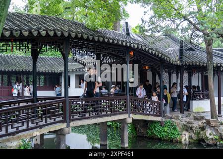 Suzhou, China - 11. Juni 2024 : Eine Holzbrücke mit komplizierten Gittergeländern überquert einen ruhigen Teich in einem üppigen chinesischen Garten. Stockfoto