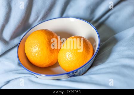 Zwei Orangen-Früchte auf einer weißen Keramikschale auf dem hellblauen Stoff im Hintergrund. Stockfoto