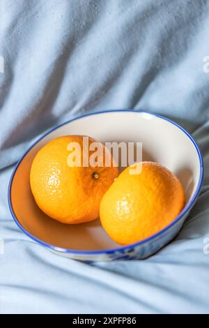 Zwei Orangen-Früchte auf einer weißen Keramikschale auf dem hellblauen Stoff im Hintergrund. Stockfoto