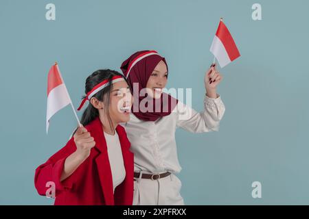 Zwei aufgeregte indonesische Frauen, die die Mini-Landflagge halten, lächelnd und unterstützend, isoliert durch blauen Hintergrund. Indonesien Unabhängigkeitstag Konzept. Stockfoto