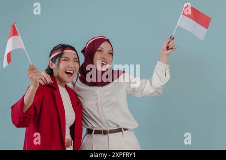 Zwei aufgeregte indonesische Frauen, die die Mini-Landflagge halten, lächelnd und unterstützend, isoliert durch blauen Hintergrund. Indonesien Unabhängigkeitstag Konzept. Stockfoto