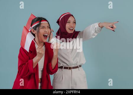 Zwei junge indonesische Frauen, die eine Mini-Landflagge halten und auf etwas zeigen, isoliert durch blauen Hintergrund. Indonesien Unabhängigkeitstag Konzept. Stockfoto