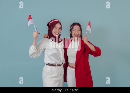 Zwei attraktive indonesische Frauen, die eine Mini-Landflagge halten, mit einem glücklichen, erfolgreichen Ausdruck, isoliert durch blauen Hintergrund. Indonesien Unabhängigkeitstag Stockfoto