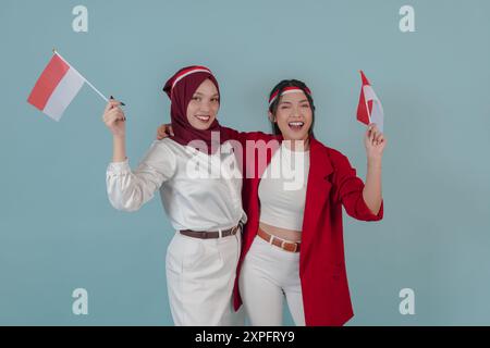 Zwei attraktive indonesische Frauen, die eine Mini-Landflagge halten, mit einem glücklichen, erfolgreichen Ausdruck, isoliert durch blauen Hintergrund. Indonesien Unabhängigkeitstag Stockfoto