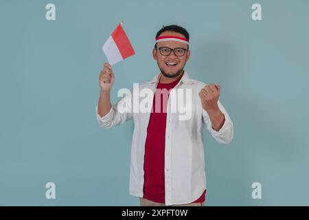 Ein lächelnder junger indonesischer Mann in einer Brille, der die Mini-Landflagge hält, während er mit einem glücklichen, erfolgreichen Ausdruck die Faust hebt. Indonesien Unabhängigkeitstag Stockfoto