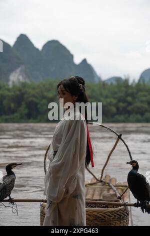 Hanfu-Frau in der Han-Dynastie kleidet sich auf einem Bambusfloß mit einem Kormoran, der Gesten mit einer Hand macht, für eine Fotosession vor der malerischen Kulisse o Stockfoto