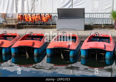 Mehrere rote Boote parken ordentlich am Wasser und reflektieren hell im Sonnenlicht, neben Schwimmwesten, die in der Nähe hängen. Stockfoto