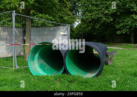 Zwei grüne Abflussrohre liegen an klaren Tagen auf dem Gras neben einer umzäunten Baustelle, umgeben von Bäumen. Stockfoto