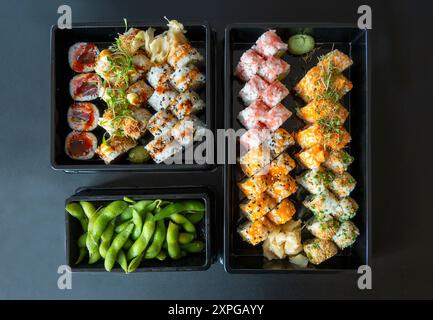 Köstliche Sushi-Brötchen nebeneinander und süße Edamame-Erbsen in schwarzen Tabletts. Draufsicht auf blauem und schwarzem Hintergrund. Stockfoto