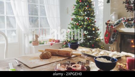 Friedlicher verschneite Weihnachtsmorgen: Aufnahme eines Tisches mit Backzutaten und Utensilien in einer dekorierten Ecke eines modernen Hauses mit Weihnachtsbaum, Kamin und Geschenken. Urlaubsvorbereitungen Stockfoto