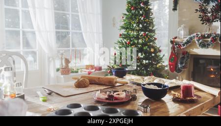 Friedlicher verschneite Weihnachtsmorgen: Tisch mit Backzutaten und Utensilien in einer dekorierten Ecke eines modernen Hauses mit Weihnachtsbaum, Kamin und Geschenken. Urlaubsvorbereitungen Stockfoto